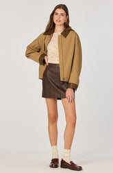 A woman is wearing a tan jacket with dark brown collar and cuffs, paired with a beige lace top. She has on a high-waisted dark brown leather mini skirt, cream-colored knit socks, and dark brown loafers. She is accessorized with gold earrings and rings.